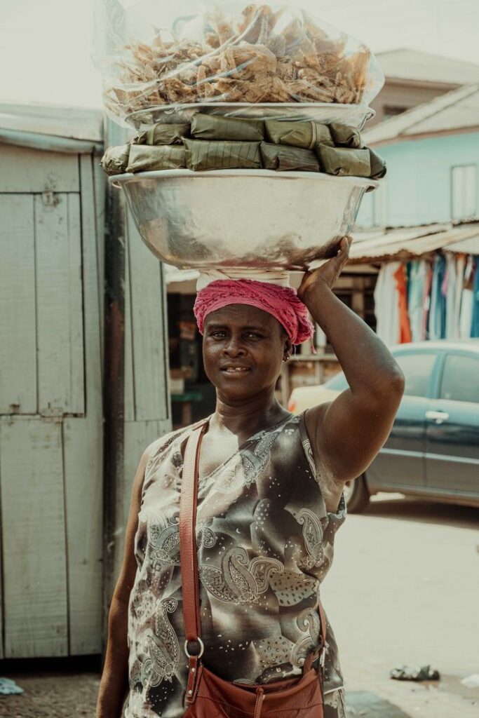 African woman balancing a bowl of goods on her head in a bustling Kumasi street.