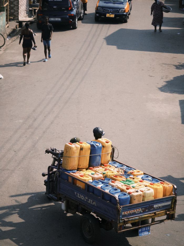 A bustling street in Ghana featuring a motor vehicle carrying colorful fuel containers and pedestrians.